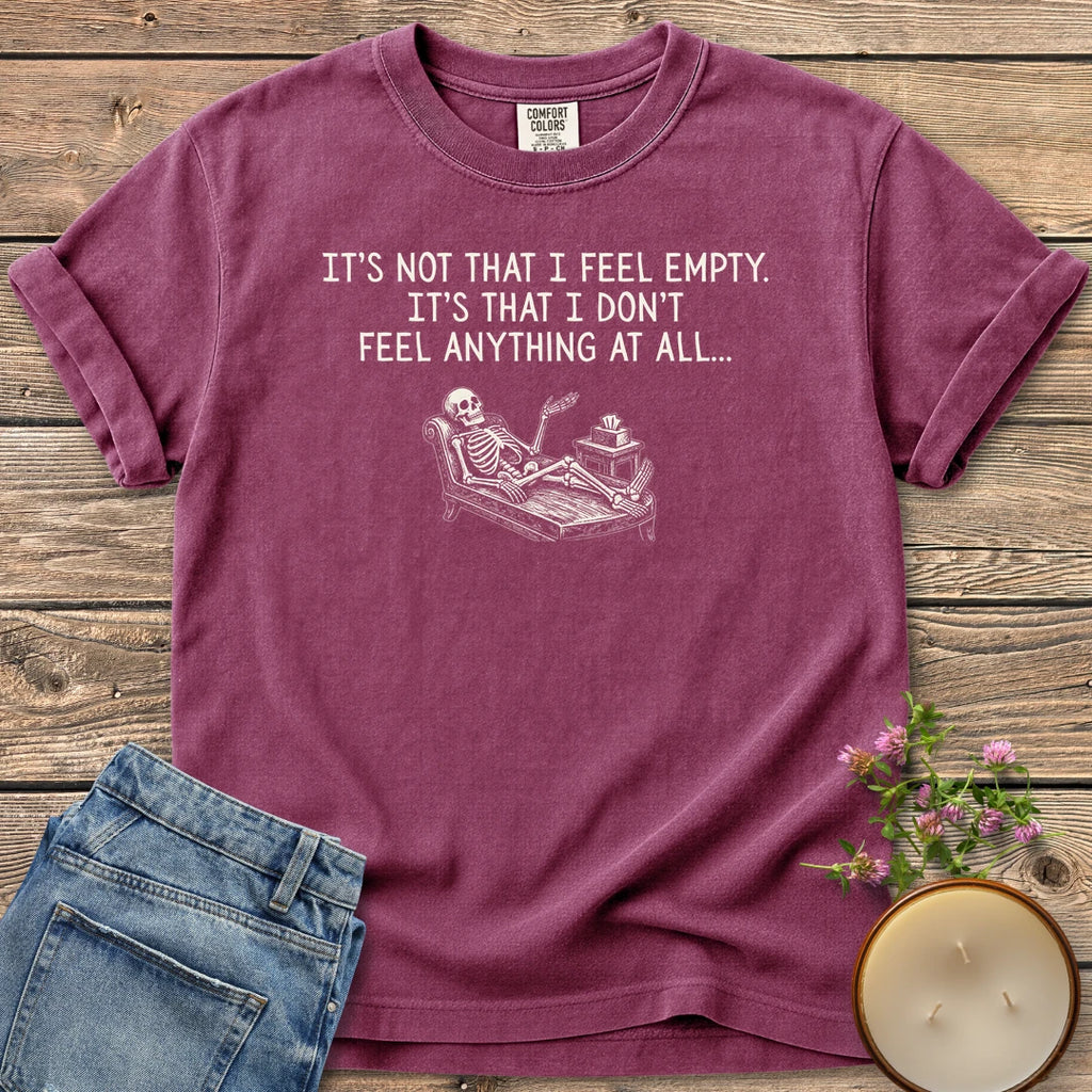 Don't Feel Anything At All t-shirt, a skeleton on a therapy couch under It's Not That I Feel Empty. It's That I Don't Feel Anything At All, on a Berry Comfort Colors tee