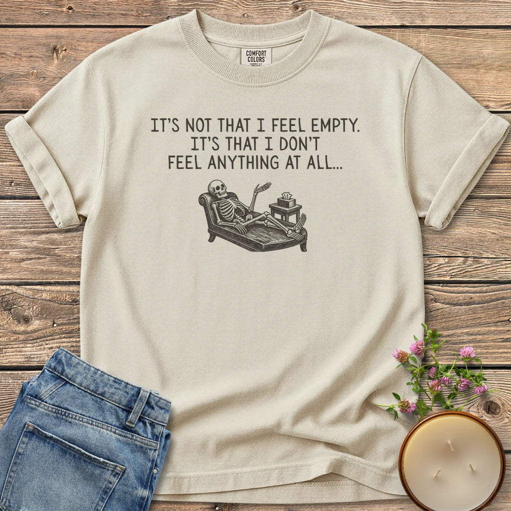 Don't Feel Anything At All t-shirt, a skeleton on a therapy couch under It's Not That I Feel Empty. It's That I Don't Feel Anything At All, on an Ivory Comfort Colors tee