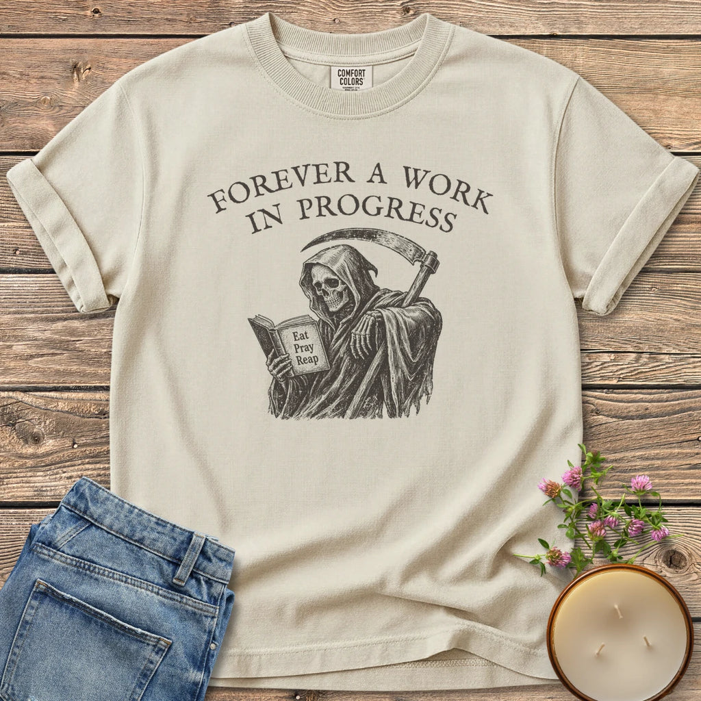 Eat Pray Reap t-shirt, a grim reaper reading a book titled Eat Pray Reap under Forever A Work In Progress, on an Ivory Comfort Colors tee