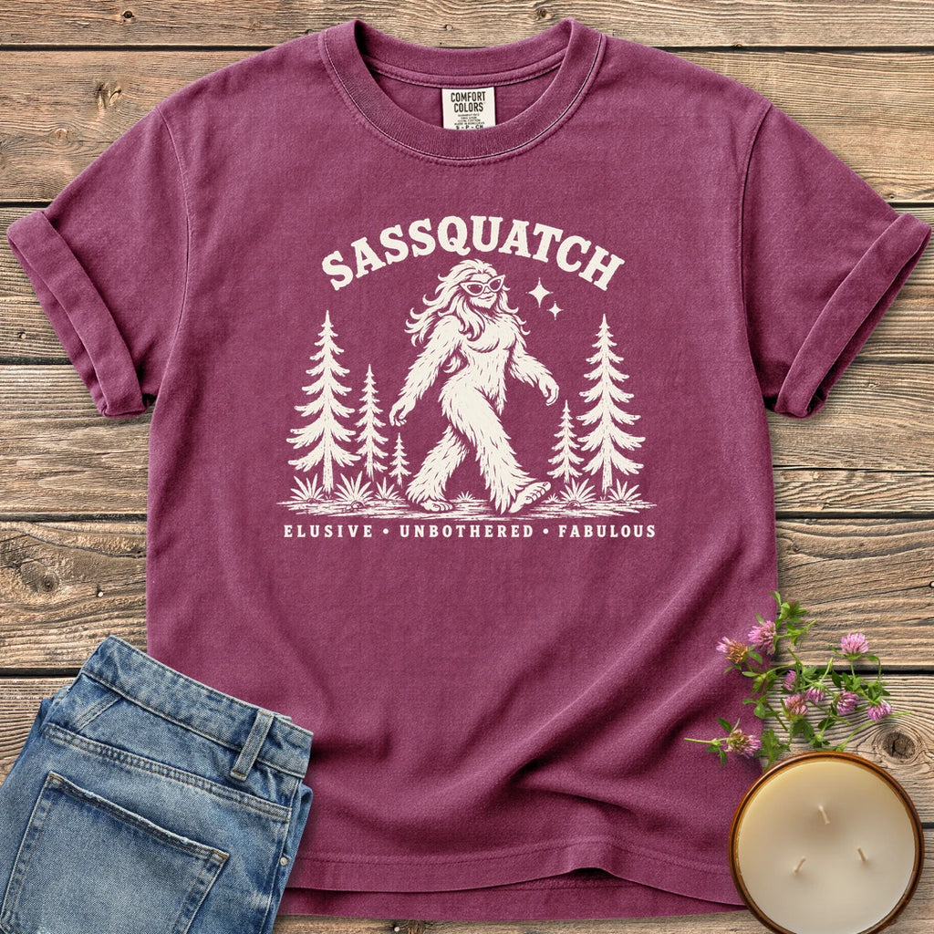 Elusive Unbothered Fabulous t-shirt, a Sassquatch in cat-eye glasses striding through pine trees above Elusive, Unbothered, Fabulous, on a Berry Comfort Colors tee