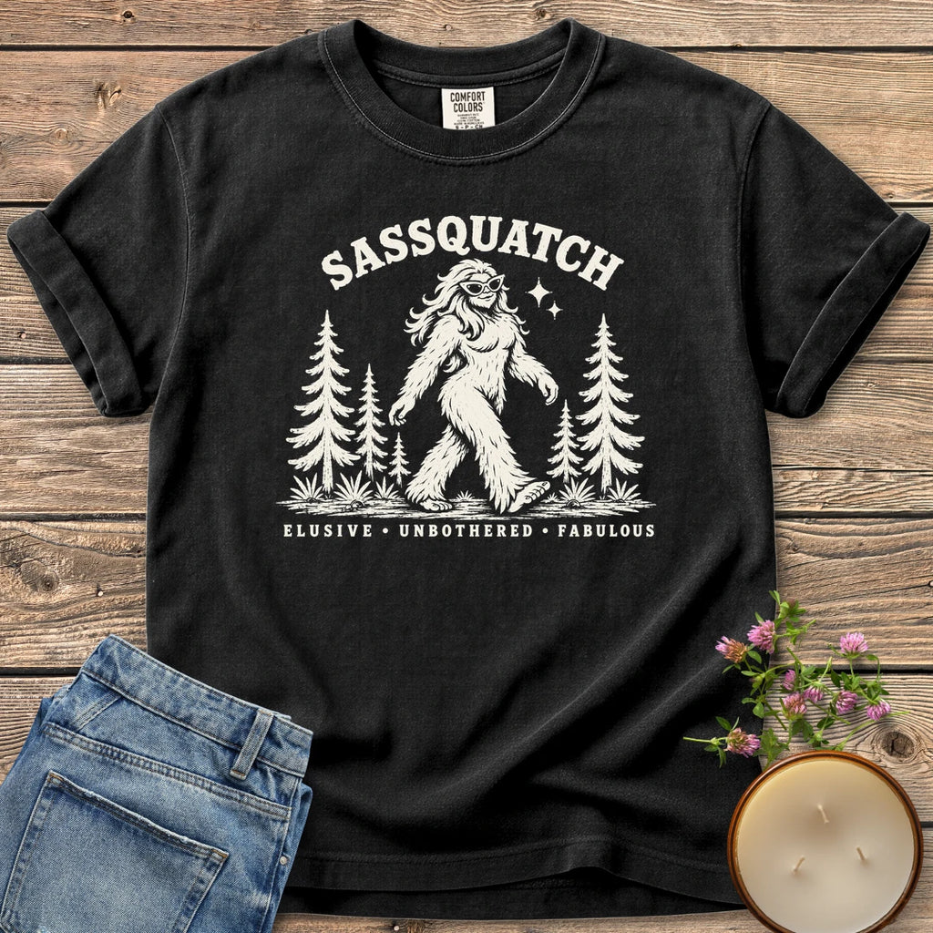 Elusive Unbothered Fabulous t-shirt, a Sassquatch in cat-eye glasses striding through pine trees above Elusive, Unbothered, Fabulous, on a Black Comfort Colors tee