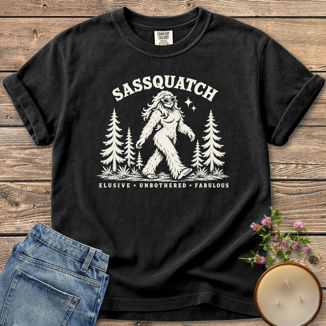 Elusive Unbothered Fabulous t-shirt, a Sassquatch in cat-eye glasses striding through pine trees above Elusive, Unbothered, Fabulous, on a Black Comfort Colors tee
