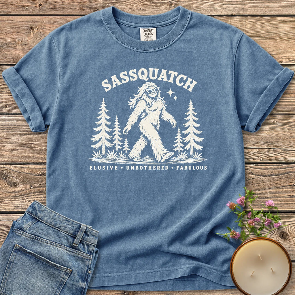 Elusive Unbothered Fabulous t-shirt, a Sassquatch in cat-eye glasses striding through pine trees above Elusive, Unbothered, Fabulous, on a Blue Jean Comfort Colors tee