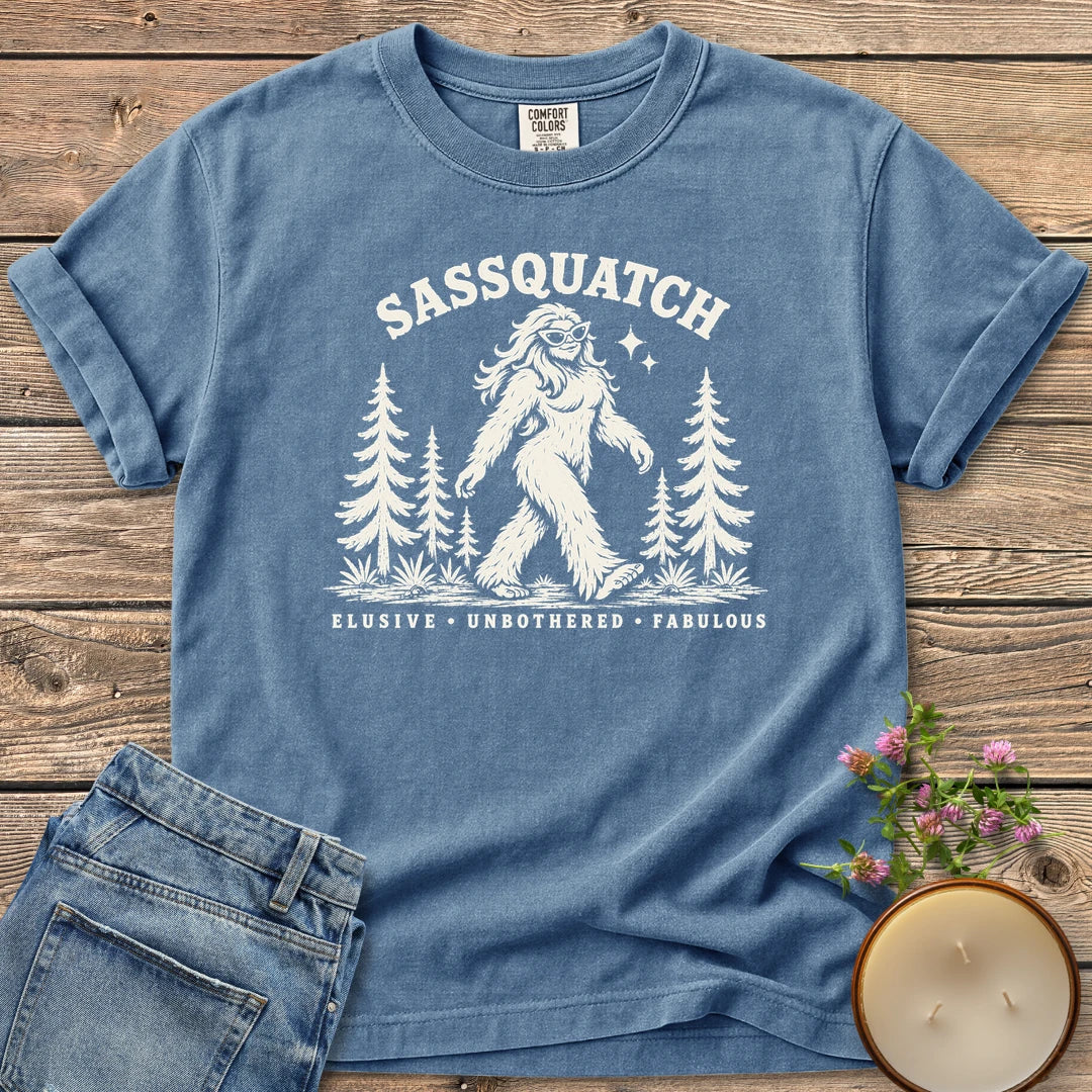 Elusive Unbothered Fabulous t-shirt, a Sassquatch in cat-eye glasses striding through pine trees above Elusive, Unbothered, Fabulous, on a Blue Jean Comfort Colors tee