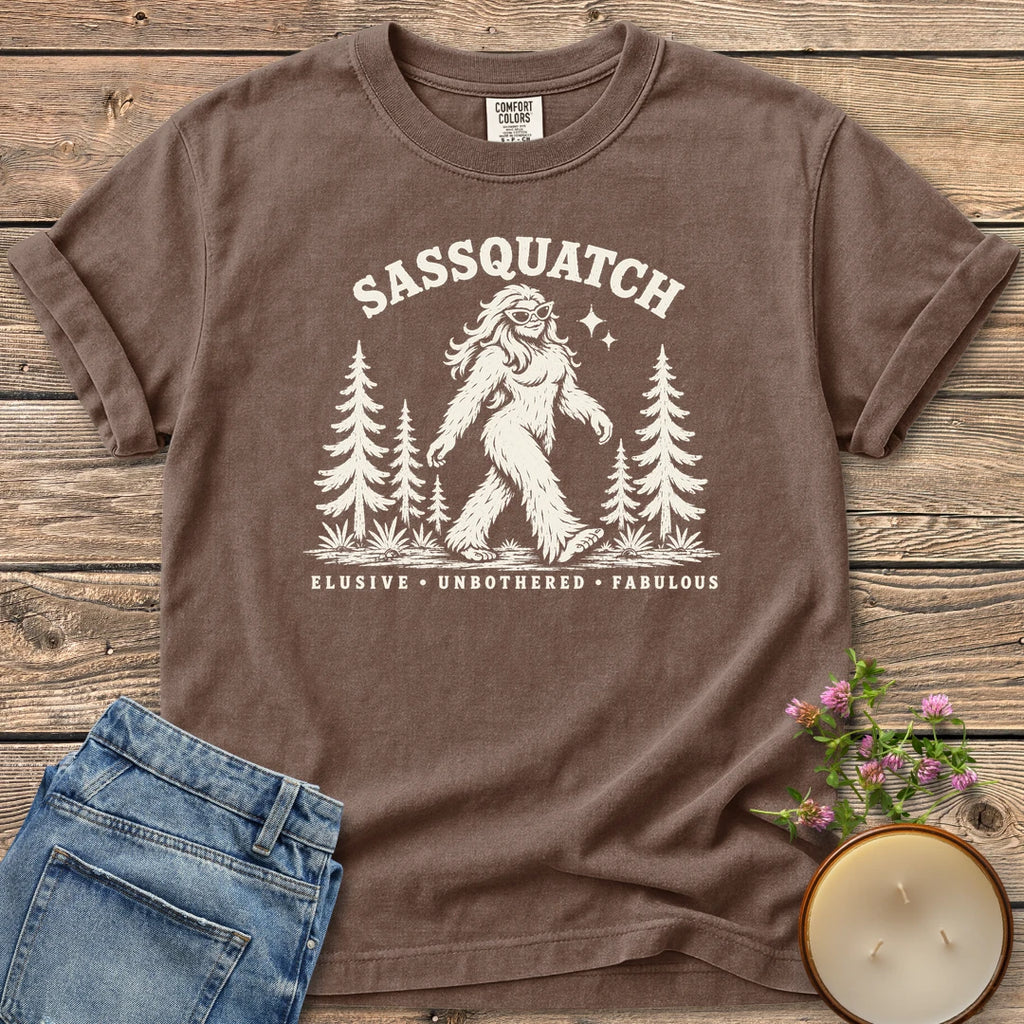 Elusive Unbothered Fabulous t-shirt, a Sassquatch in cat-eye glasses striding through pine trees above Elusive, Unbothered, Fabulous, on an Espresso Comfort Colors tee