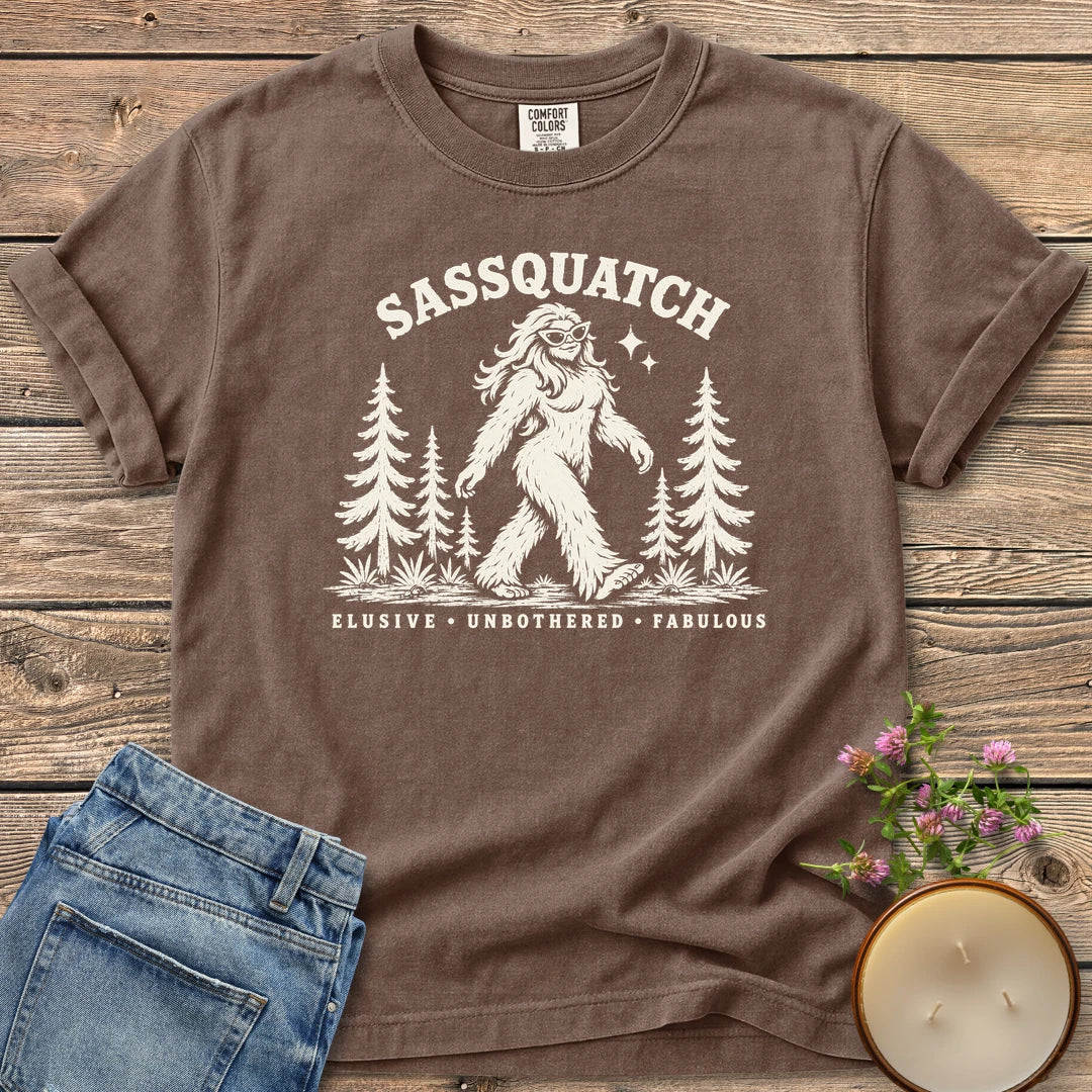 Elusive Unbothered Fabulous t-shirt, a Sassquatch in cat-eye glasses striding through pine trees above Elusive, Unbothered, Fabulous, on an Espresso Comfort Colors tee