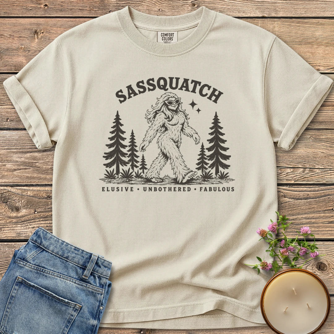 Elusive Unbothered Fabulous t-shirt, a Sassquatch in cat-eye glasses striding through pine trees above Elusive, Unbothered, Fabulous, on an Ivory Comfort Colors tee