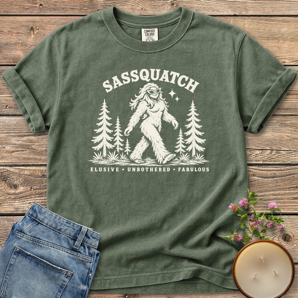 Elusive Unbothered Fabulous t-shirt, a Sassquatch in cat-eye glasses striding through pine trees above Elusive, Unbothered, Fabulous, on a Moss Comfort Colors tee