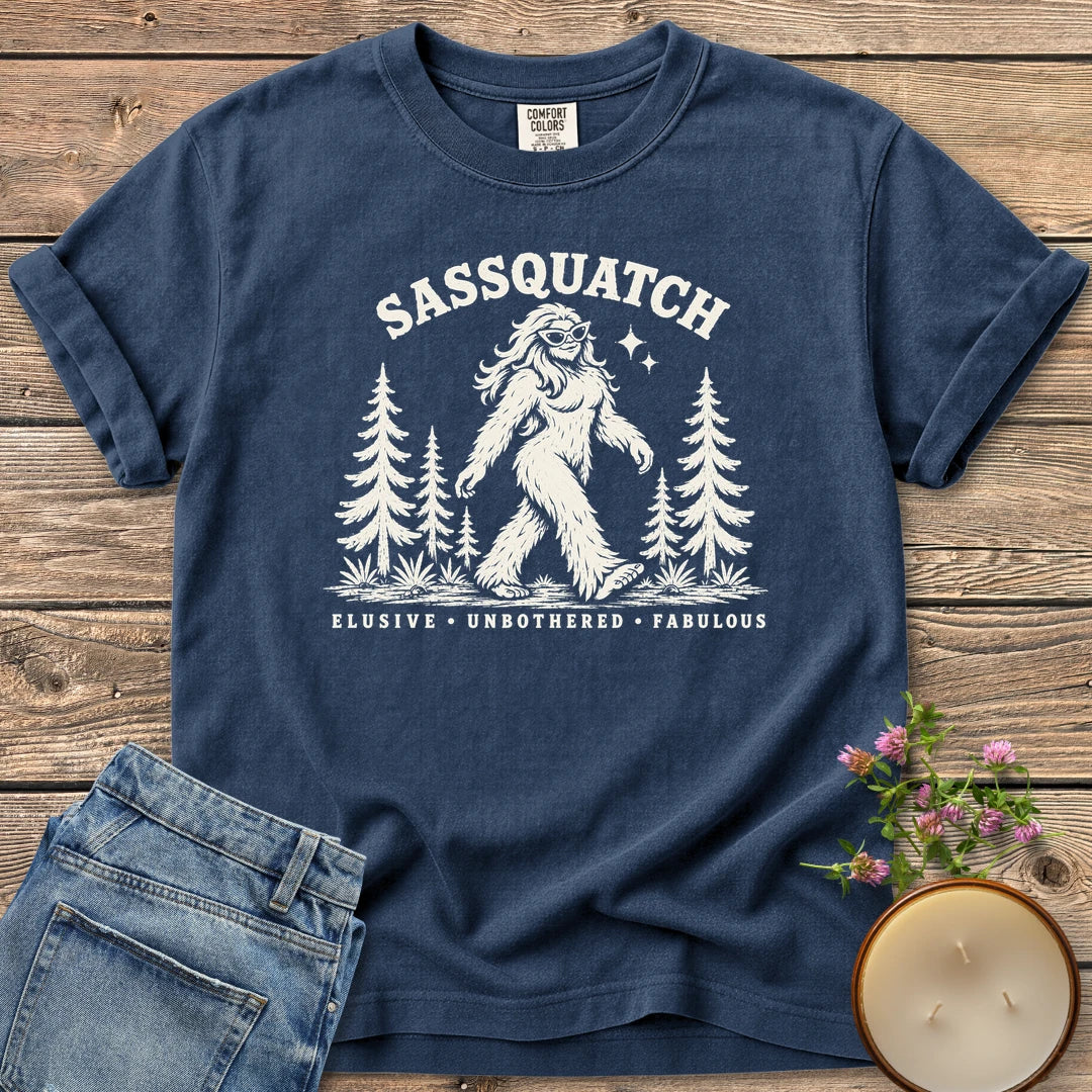 Elusive Unbothered Fabulous t-shirt, a Sassquatch in cat-eye glasses striding through pine trees above Elusive, Unbothered, Fabulous, on a Navy Comfort Colors tee