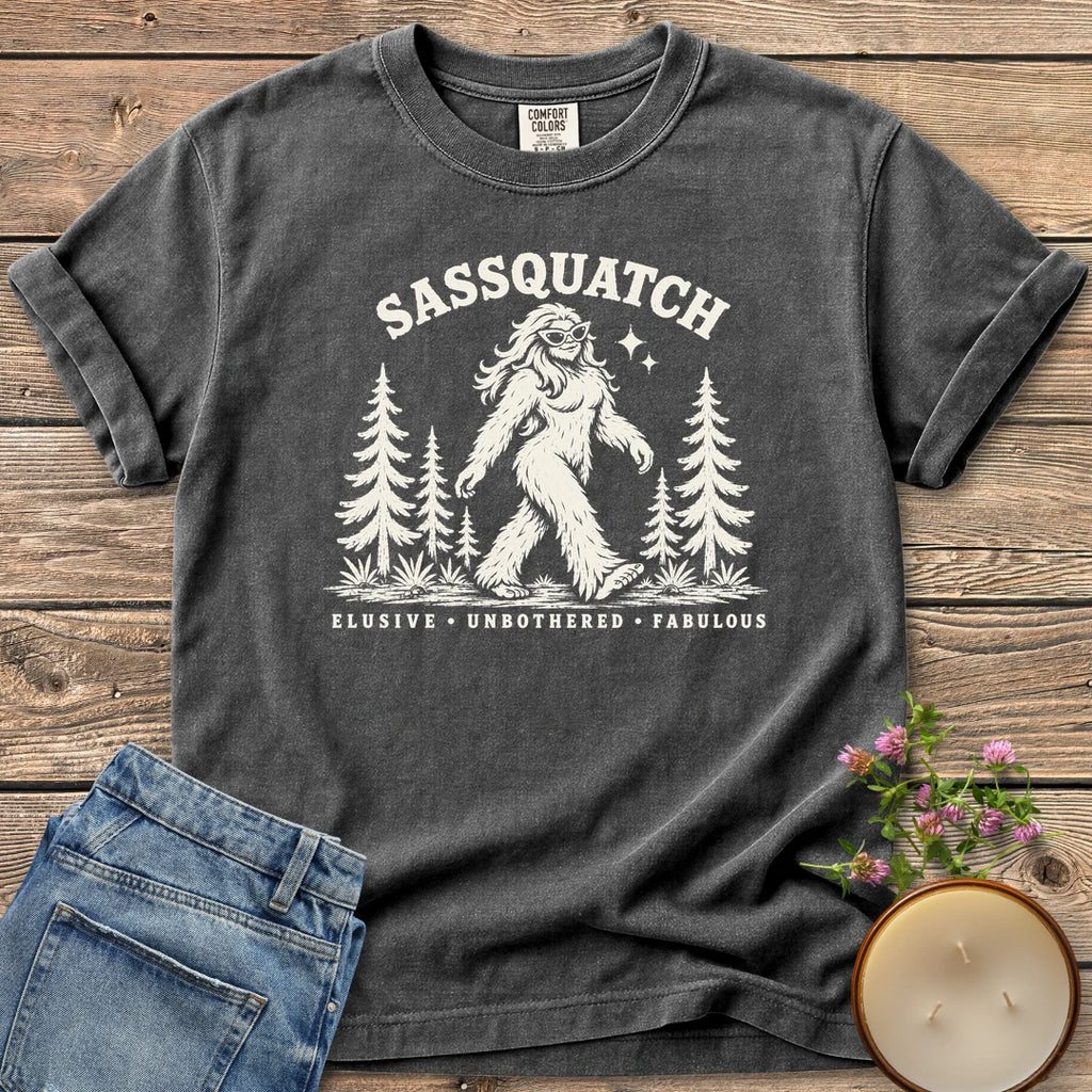 Elusive Unbothered Fabulous t-shirt, a Sassquatch in cat-eye glasses striding through pine trees above Elusive, Unbothered, Fabulous, on a Pepper Comfort Colors tee