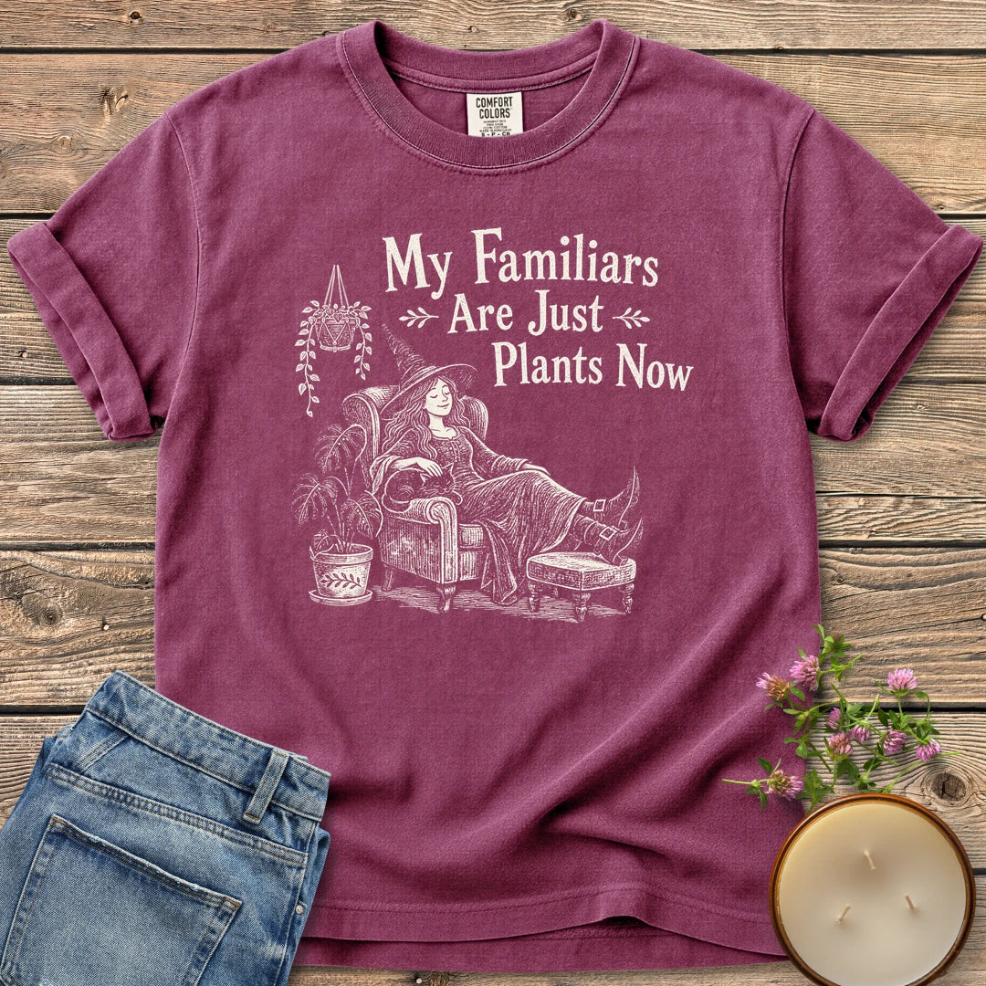 Familiars Are Just Plants Now t-shirt, a relaxed witch lounging in an armchair among houseplants under My Familiars Are Just Plants Now, on a Berry Comfort Colors tee
