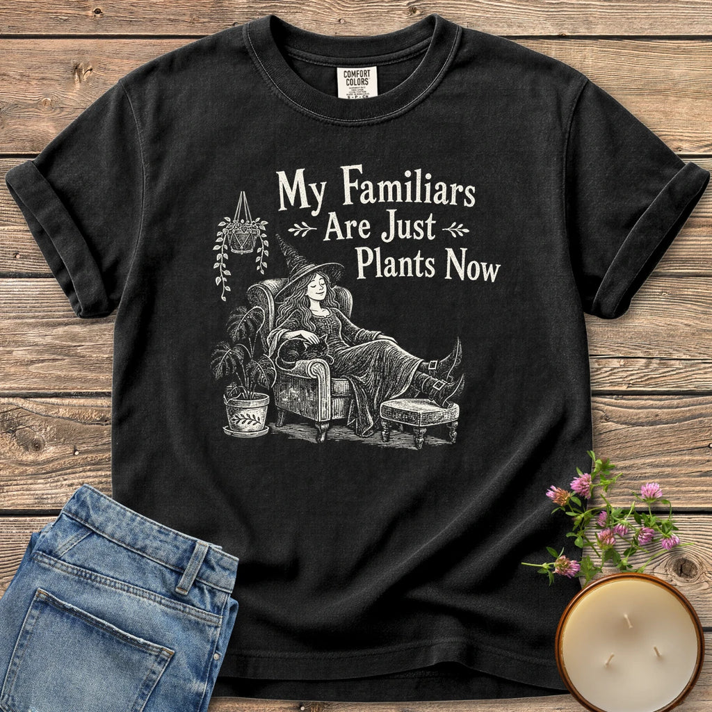Familiars Are Just Plants Now t-shirt, a relaxed witch lounging in an armchair among houseplants under My Familiars Are Just Plants Now, on a Black Comfort Colors tee