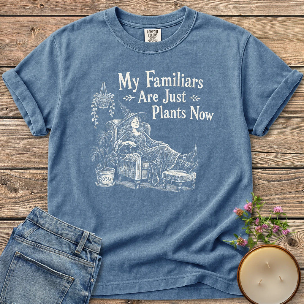 Familiars Are Just Plants Now t-shirt, a relaxed witch lounging in an armchair among houseplants under My Familiars Are Just Plants Now, on a Blue Jean Comfort Colors tee