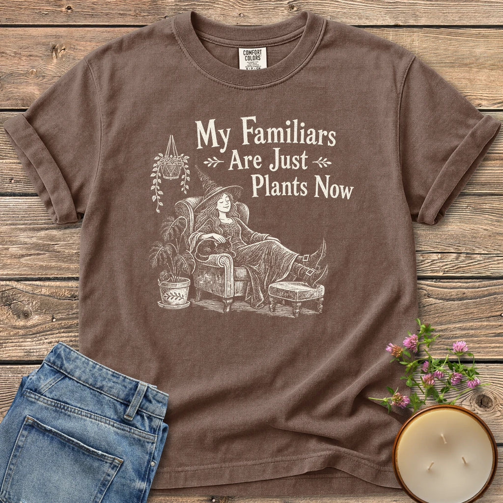 Familiars Are Just Plants Now t-shirt, a relaxed witch lounging in an armchair among houseplants under My Familiars Are Just Plants Now, on an Espresso Comfort Colors tee