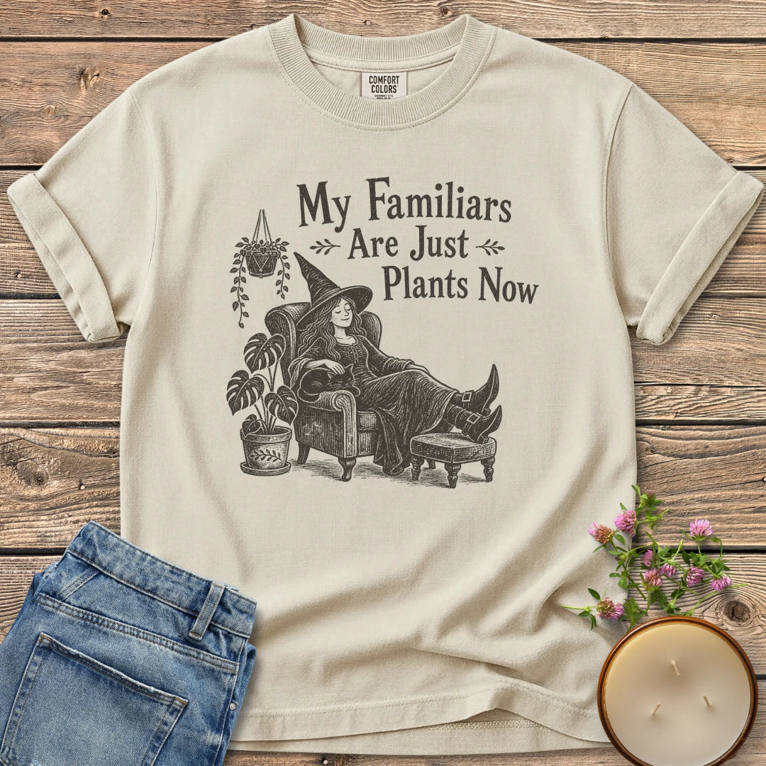 Familiars Are Just Plants Now t-shirt, a relaxed witch lounging in an armchair among houseplants under My Familiars Are Just Plants Now, on an Ivory Comfort Colors tee