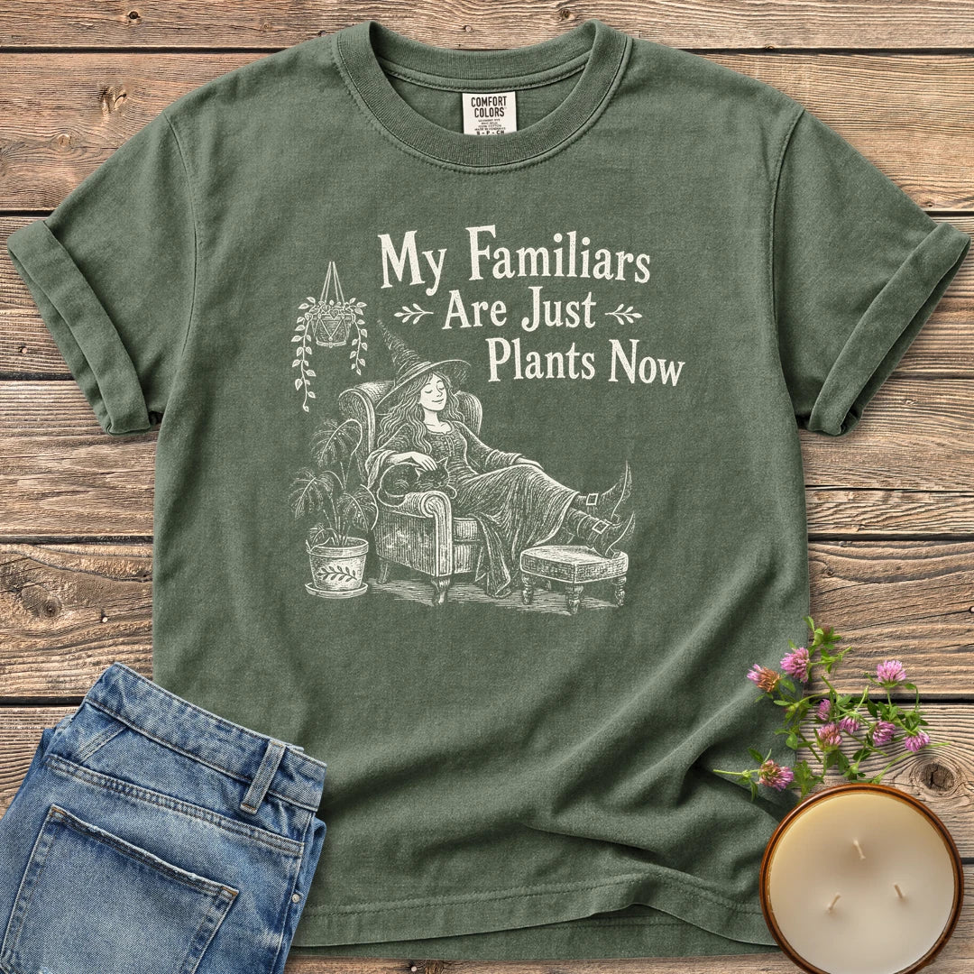 Familiars Are Just Plants Now t-shirt, a relaxed witch lounging in an armchair among houseplants under My Familiars Are Just Plants Now, on a Moss Comfort Colors tee