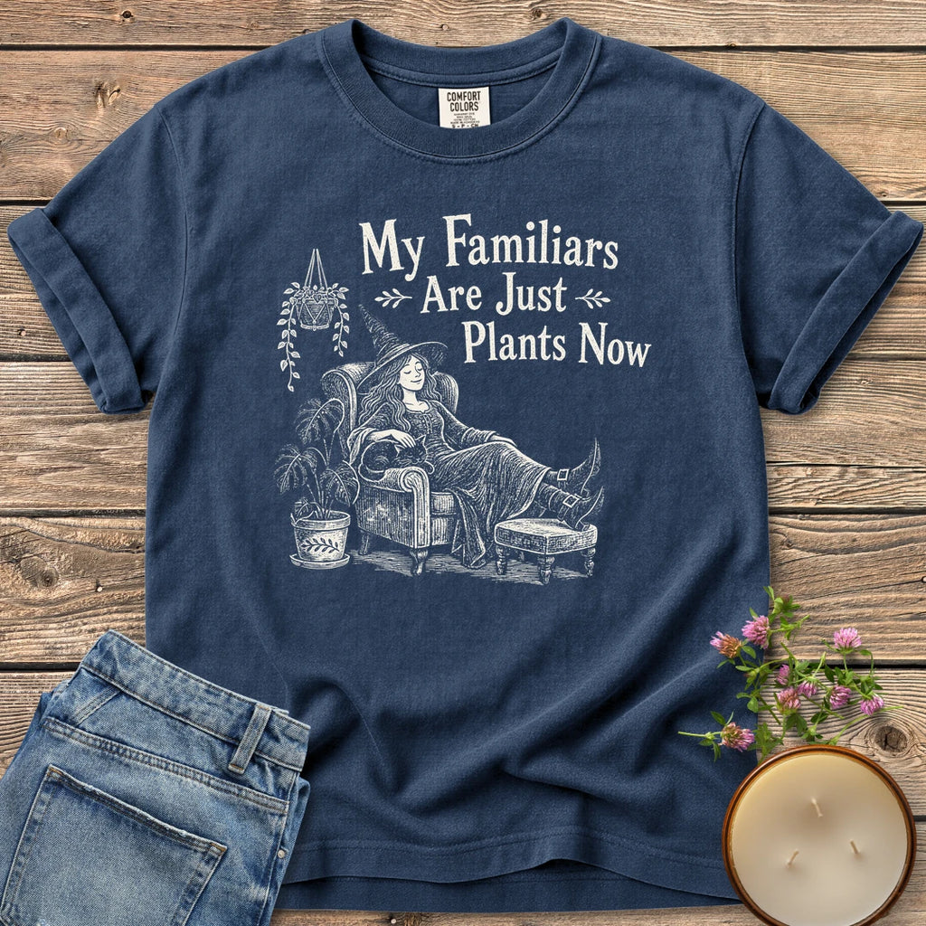 Familiars Are Just Plants Now t-shirt, a relaxed witch lounging in an armchair among houseplants under My Familiars Are Just Plants Now, on a Navy Comfort Colors tee