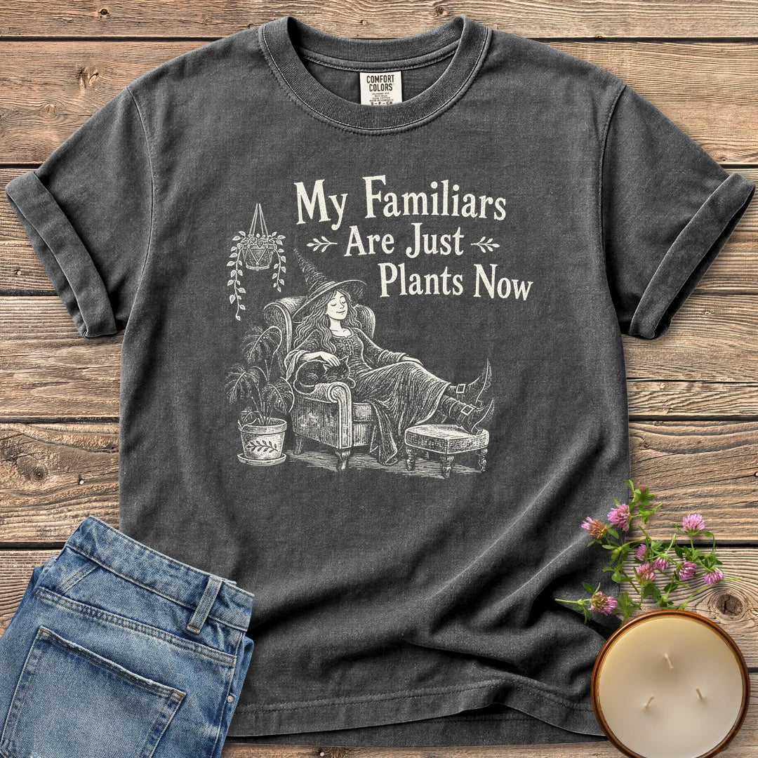 Familiars Are Just Plants Now t-shirt, a relaxed witch lounging in an armchair among houseplants under My Familiars Are Just Plants Now, on a Pepper Comfort Colors tee
