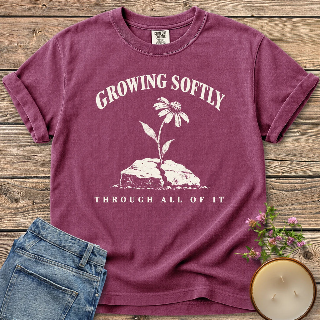 Growing Softly Through It All t-shirt, Growing Softly above a flower breaking through a cracked rock and Through All Of It, on a Berry Comfort Colors tee
