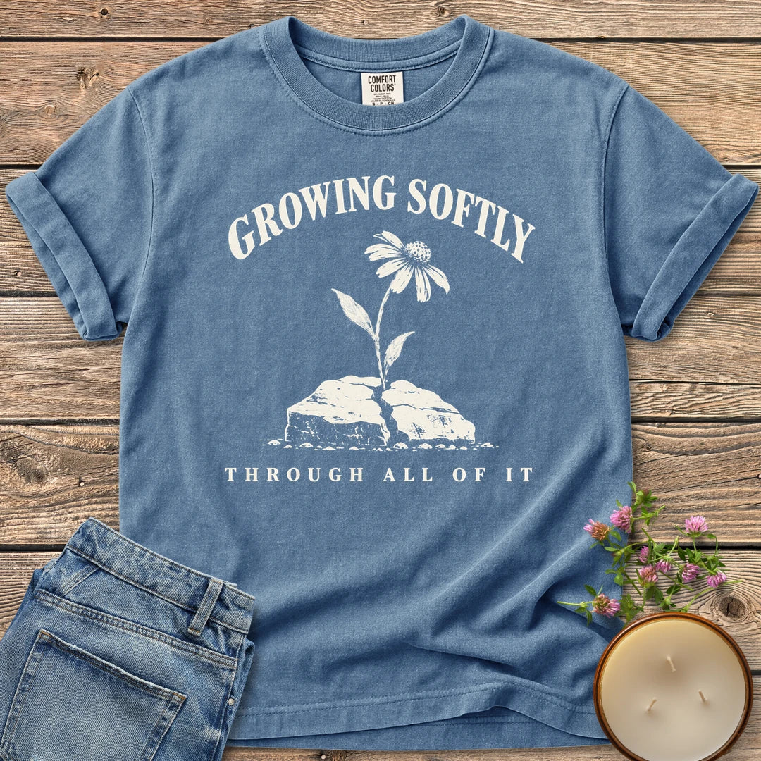 Growing Softly Through It All t-shirt, Growing Softly above a flower breaking through a cracked rock and Through All Of It, on a Blue Jean Comfort Colors tee