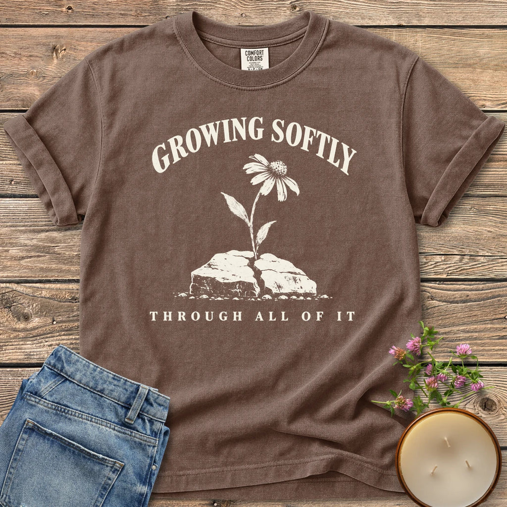 Growing Softly Through It All t-shirt, Growing Softly above a flower breaking through a cracked rock and Through All Of It, on an Espresso Comfort Colors tee