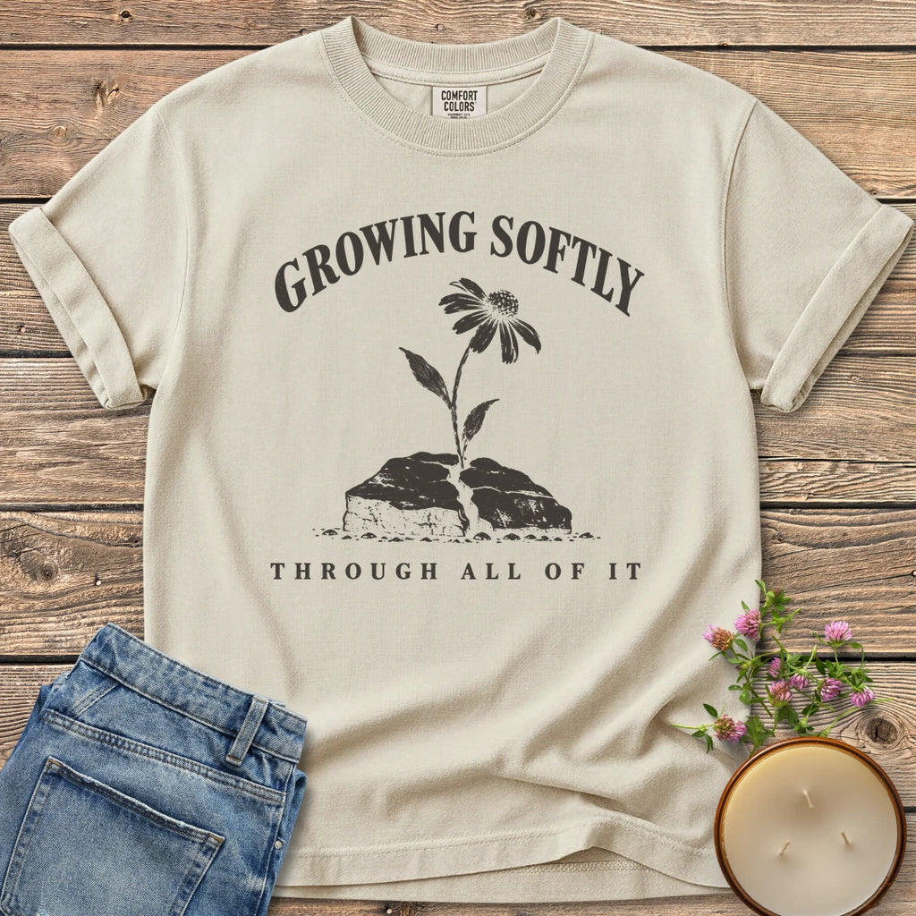 Growing Softly Through It All t-shirt, Growing Softly above a flower breaking through a cracked rock and Through All Of It, on an Ivory Comfort Colors tee
