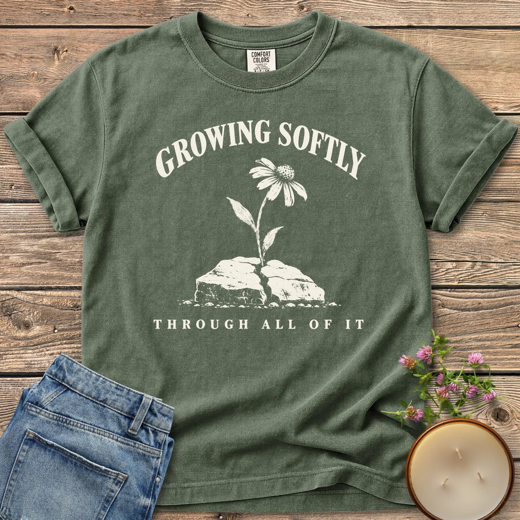 Growing Softly Through It All t-shirt, Growing Softly above a flower breaking through a cracked rock and Through All Of It, on a Moss Comfort Colors tee