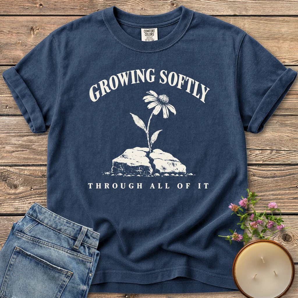 Growing Softly Through It All t-shirt, Growing Softly above a flower breaking through a cracked rock and Through All Of It, on a Navy Comfort Colors tee