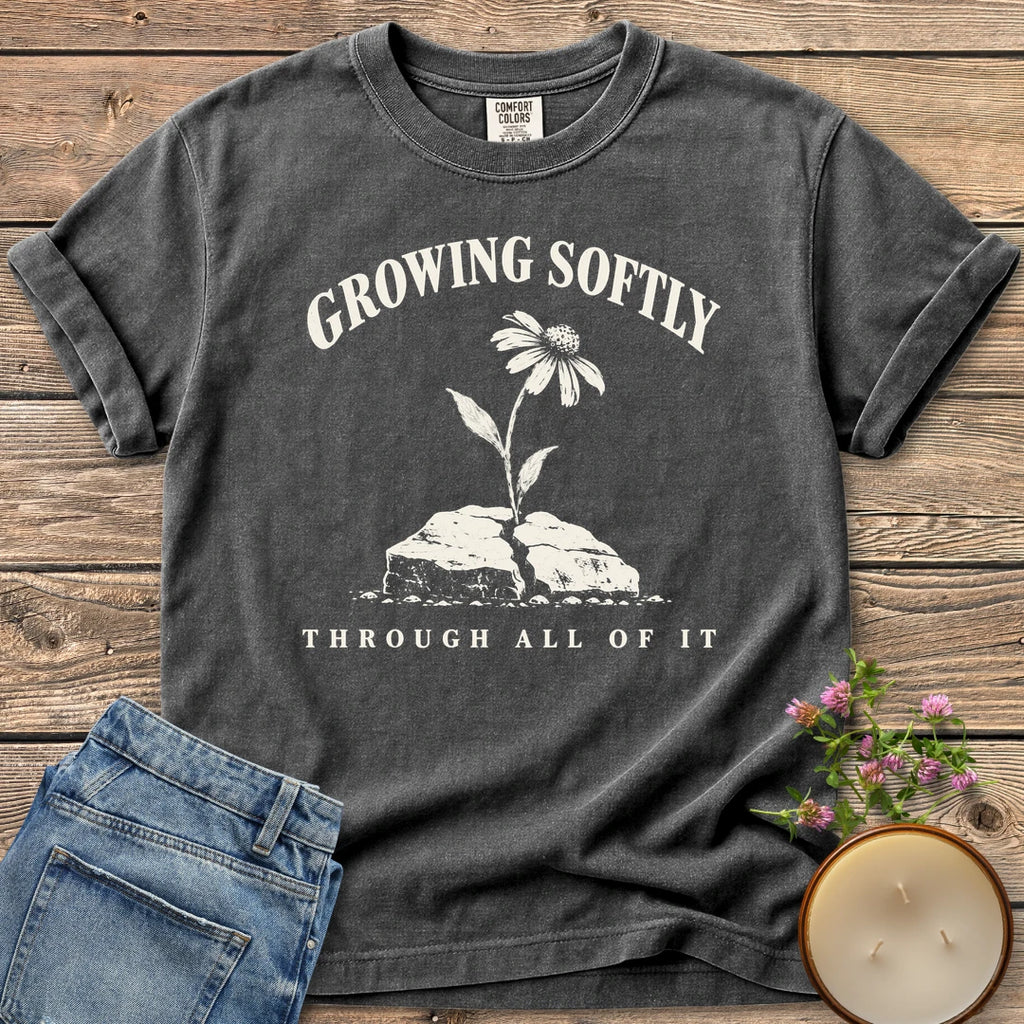 Growing Softly Through It All t-shirt, Growing Softly above a flower breaking through a cracked rock and Through All Of It, on a Pepper Comfort Colors tee