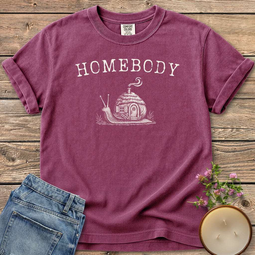 Homebody t-shirt, a snail carrying a tiny cottage with a smoking chimney under Homebody, on a Berry Comfort Colors tee