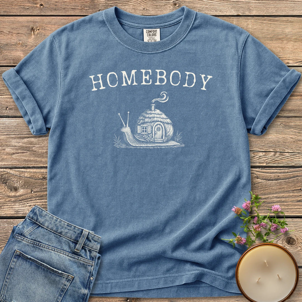 Homebody t-shirt, a snail carrying a tiny cottage with a smoking chimney under Homebody, on a Blue Jean Comfort Colors tee