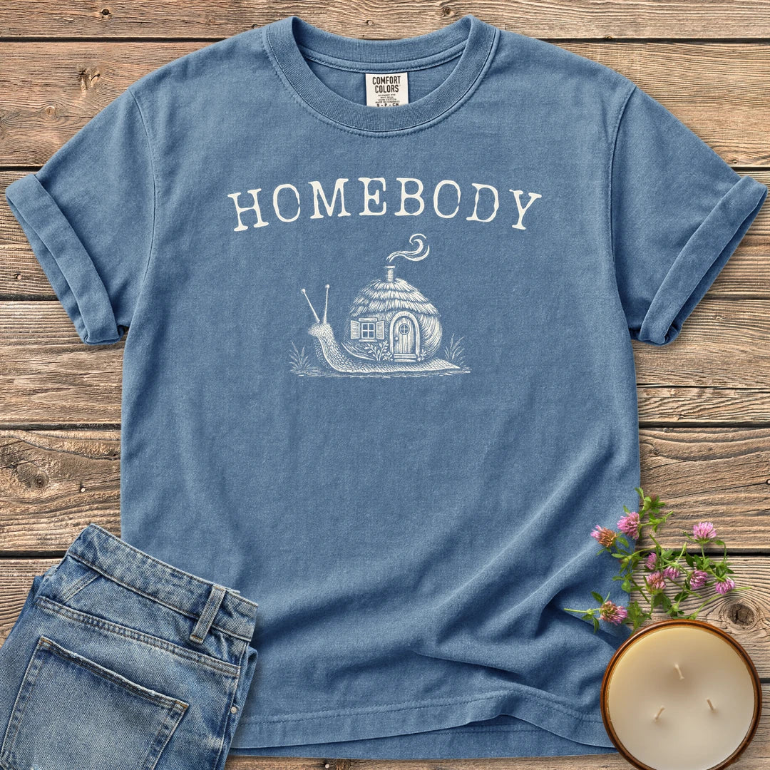 Homebody t-shirt, a snail carrying a tiny cottage with a smoking chimney under Homebody, on a Blue Jean Comfort Colors tee