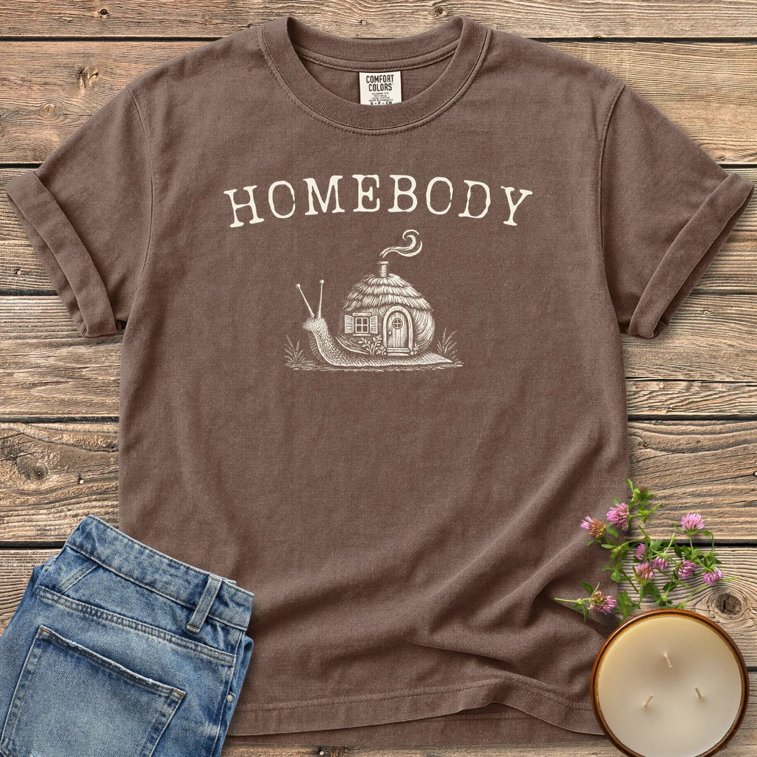 Homebody t-shirt, a snail carrying a tiny cottage with a smoking chimney under Homebody, on an Espresso Comfort Colors tee