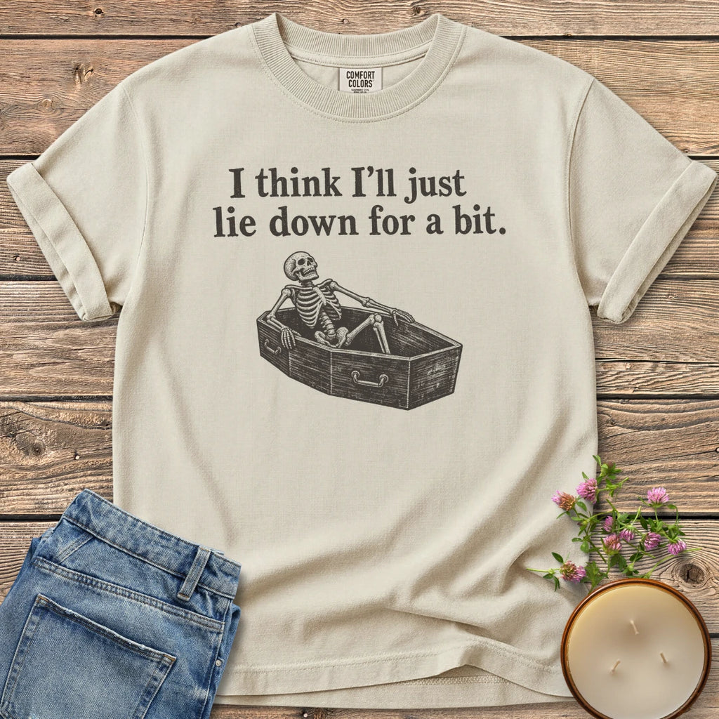 Lie Down For a Bit t-shirt, a skeleton lying in a coffin under I think I'll just lie down for a bit, on an Ivory Comfort Colors tee