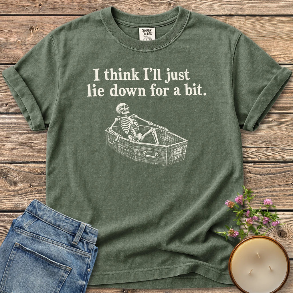 Lie Down For a Bit t-shirt, a skeleton lying in a coffin under I think I'll just lie down for a bit, on a Moss Comfort Colors tee