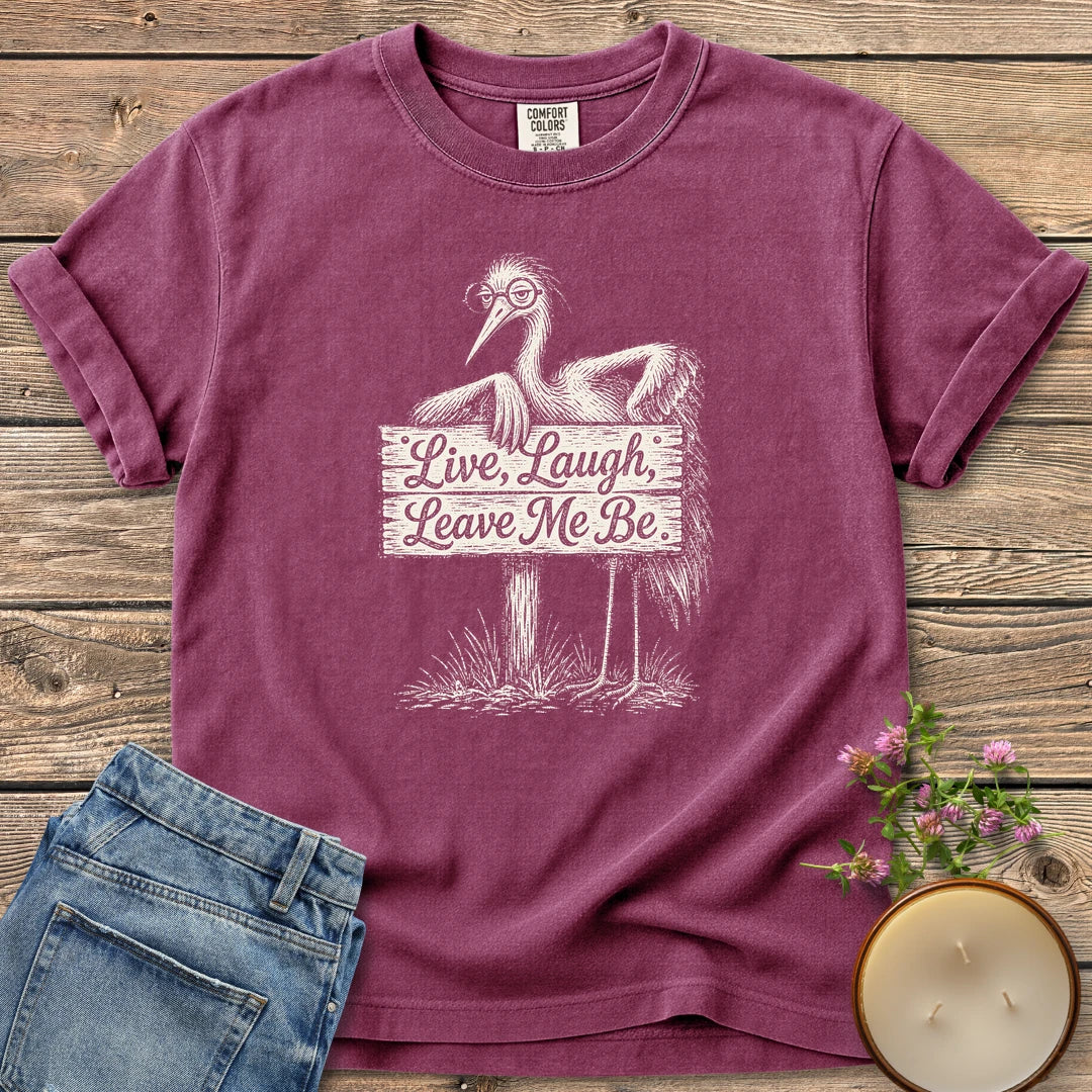 Live Laugh Leave Me Be t-shirt, a grumpy heron in glasses leaning on a sign that reads Live, Laugh, Leave Me Be, on a Berry Comfort Colors tee