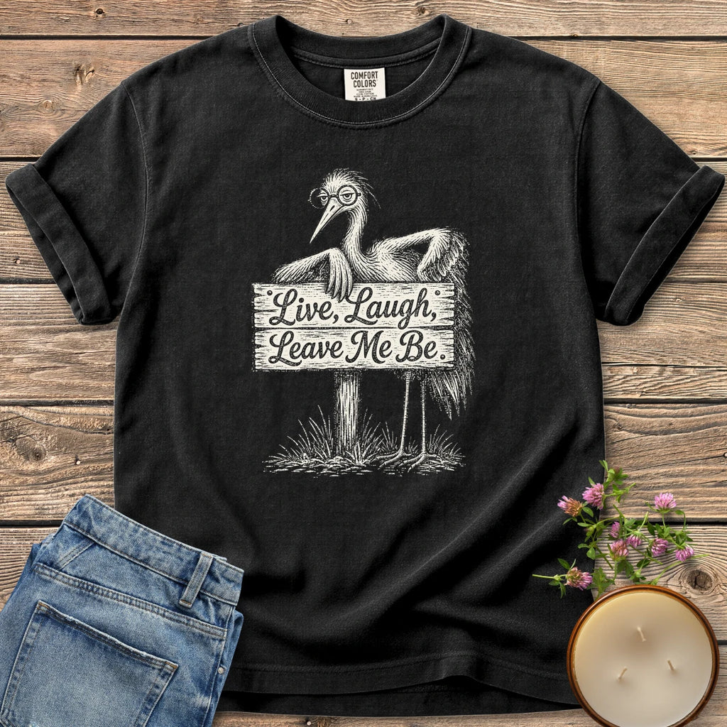 Live Laugh Leave Me Be t-shirt, a grumpy heron in glasses leaning on a sign that reads Live, Laugh, Leave Me Be, on a Black Comfort Colors tee