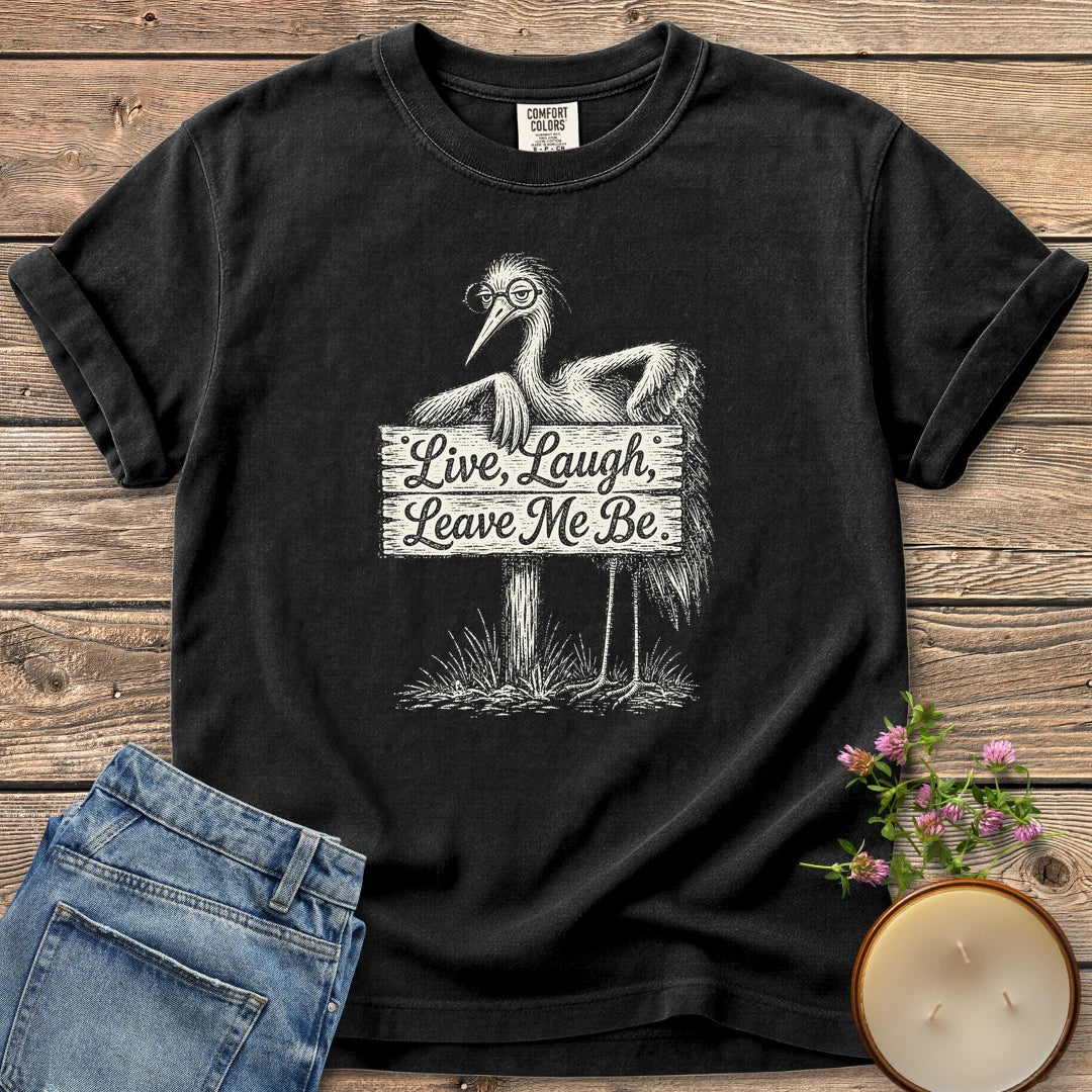 Live Laugh Leave Me Be t-shirt, a grumpy heron in glasses leaning on a sign that reads Live, Laugh, Leave Me Be, on a Black Comfort Colors tee