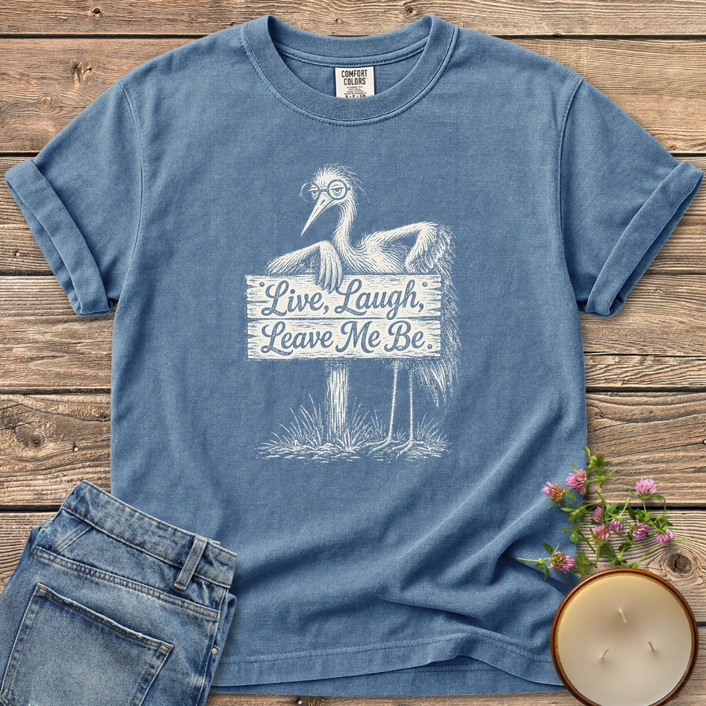 Live Laugh Leave Me Be t-shirt, a grumpy heron in glasses leaning on a sign that reads Live, Laugh, Leave Me Be, on a Blue Jean Comfort Colors tee