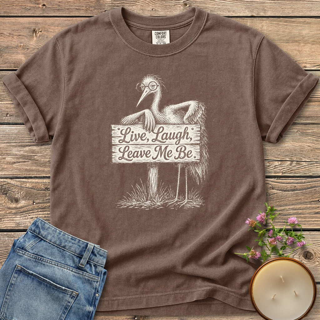 Live Laugh Leave Me Be t-shirt, a grumpy heron in glasses leaning on a sign that reads Live, Laugh, Leave Me Be, on an Espresso Comfort Colors tee