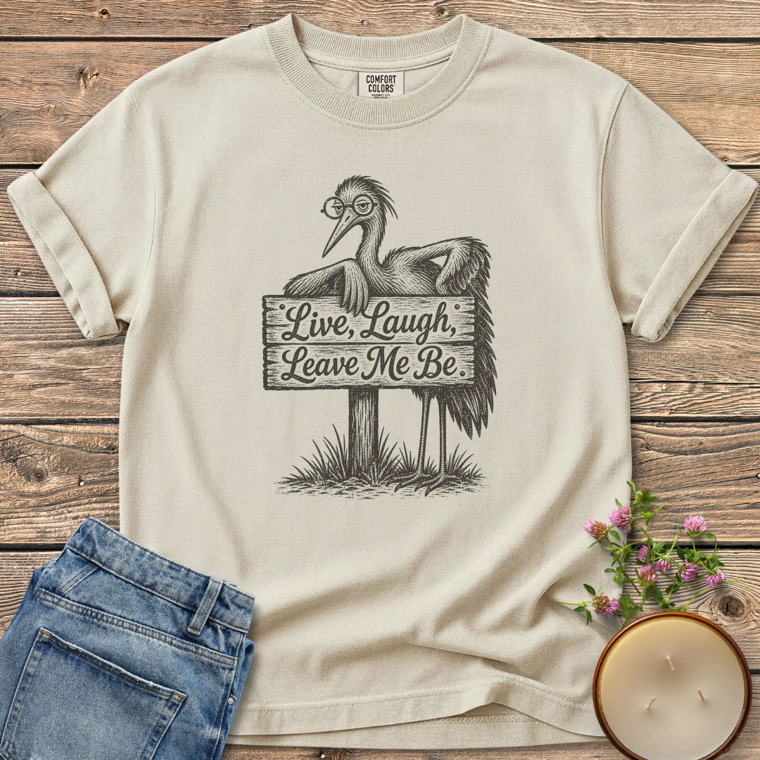 Live Laugh Leave Me Be t-shirt, a grumpy heron in glasses leaning on a sign that reads Live, Laugh, Leave Me Be, on an Ivory Comfort Colors tee