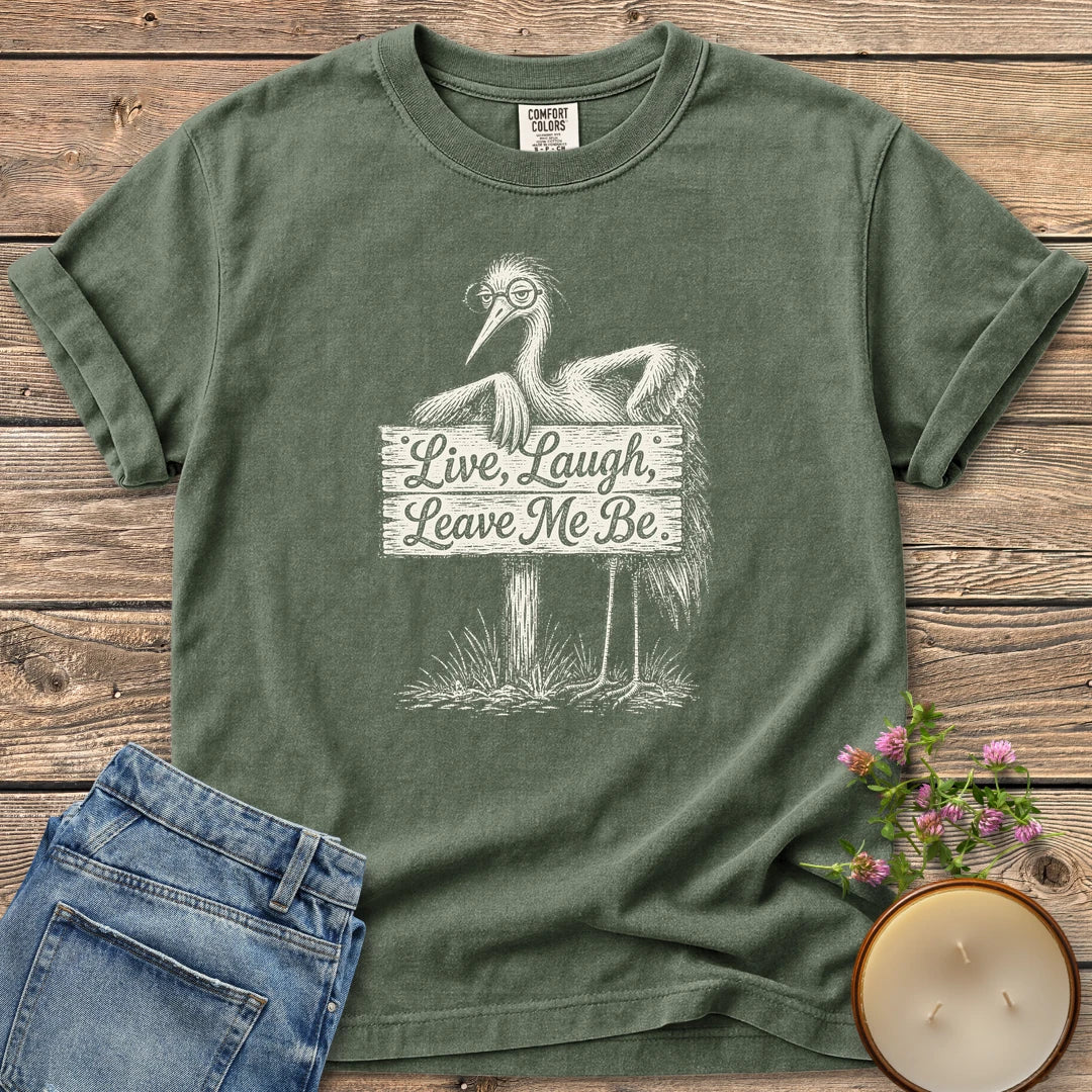 Live Laugh Leave Me Be t-shirt, a grumpy heron in glasses leaning on a sign that reads Live, Laugh, Leave Me Be, on a Moss Comfort Colors tee