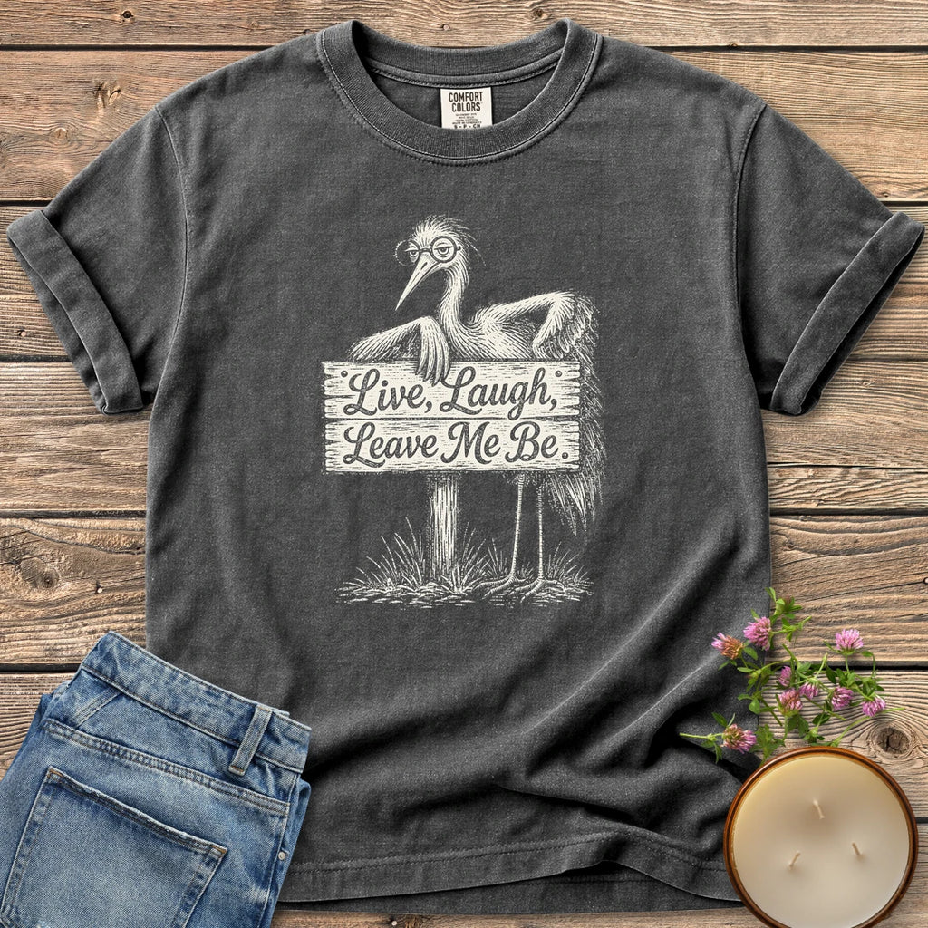 Live Laugh Leave Me Be t-shirt, a grumpy heron in glasses leaning on a sign that reads Live, Laugh, Leave Me Be, on a Pepper Comfort Colors tee