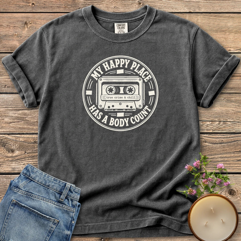 My Happy Place Body Count t-shirt, a round badge with a cassette tape reading My Happy Place Has A Body Count and true crime & chill, on a Pepper Comfort Colors tee