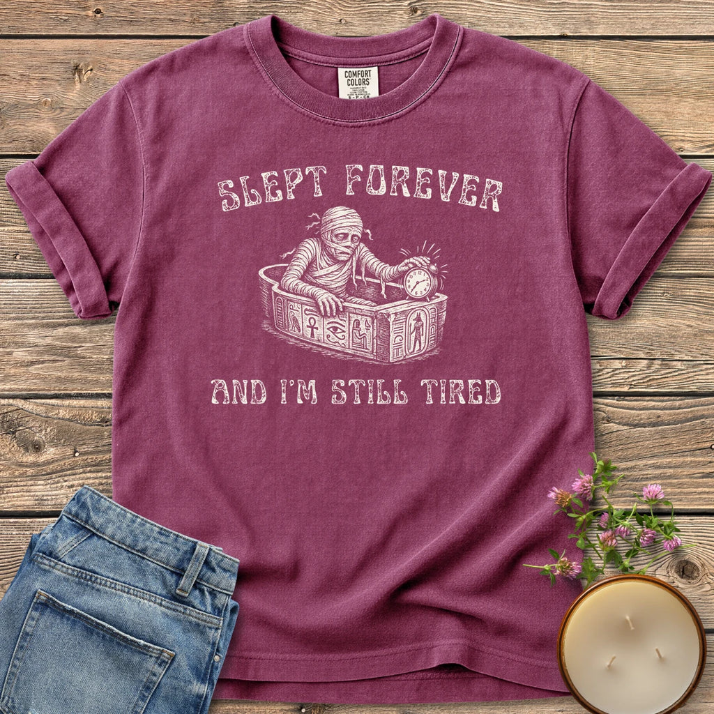 Slept Forever Still Tired t-shirt, a mummy rising from a sarcophagus hitting an alarm clock between Slept Forever and And I'm Still Tired, on a Berry Comfort Colors tee