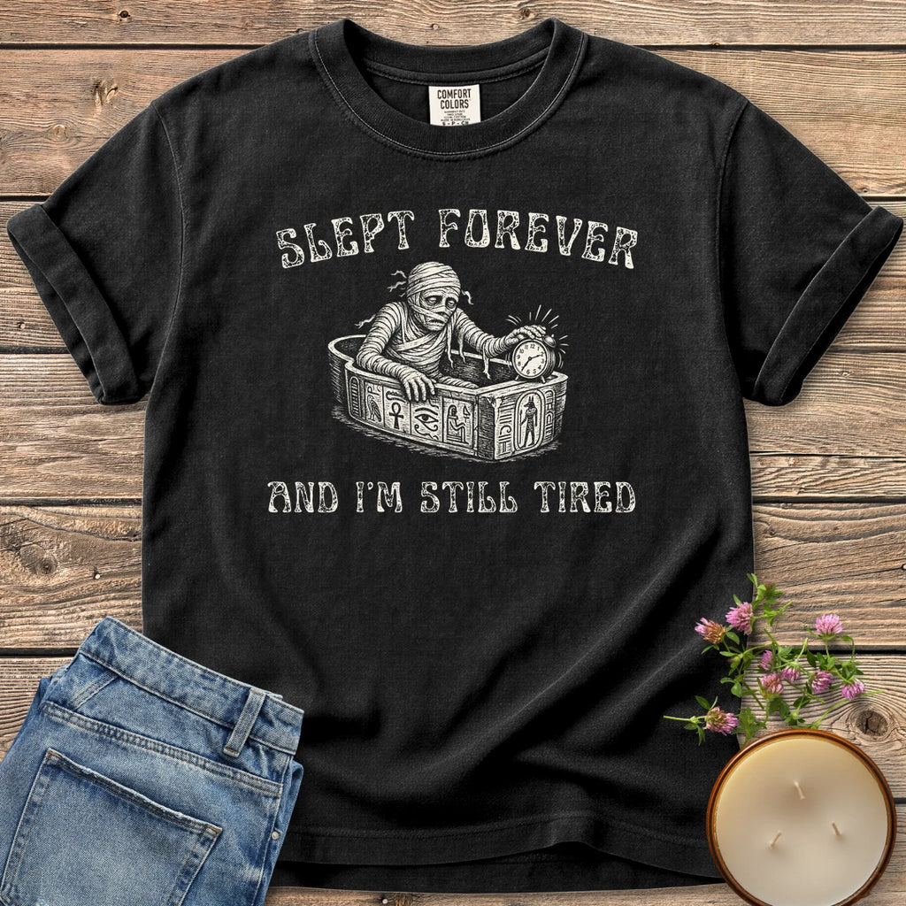 Slept Forever Still Tired t-shirt, a mummy rising from a sarcophagus hitting an alarm clock between Slept Forever and And I'm Still Tired, on a Black Comfort Colors tee