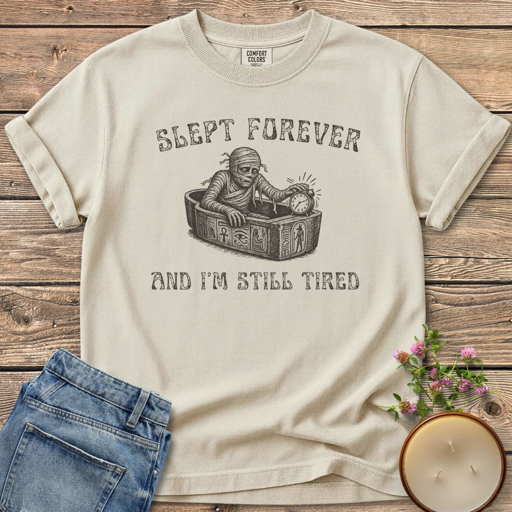 Slept Forever Still Tired t-shirt, a mummy rising from a sarcophagus hitting an alarm clock between Slept Forever and And I'm Still Tired, on an Ivory Comfort Colors tee