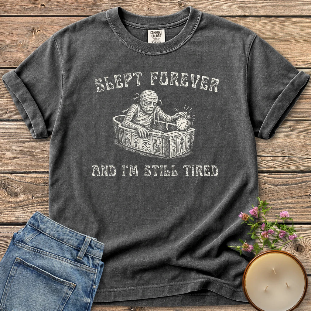 Slept Forever Still Tired t-shirt, a mummy rising from a sarcophagus hitting an alarm clock between Slept Forever and And I'm Still Tired, on a Pepper Comfort Colors tee