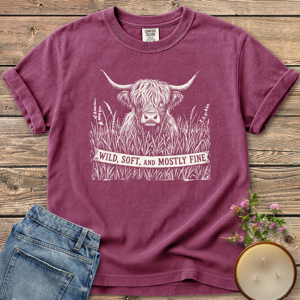 Wild Soft And Mostly Fine t-shirt, a shaggy highland cow in tall grass with a banner reading Wild, Soft, and Mostly Fine, on a Berry Comfort Colors tee